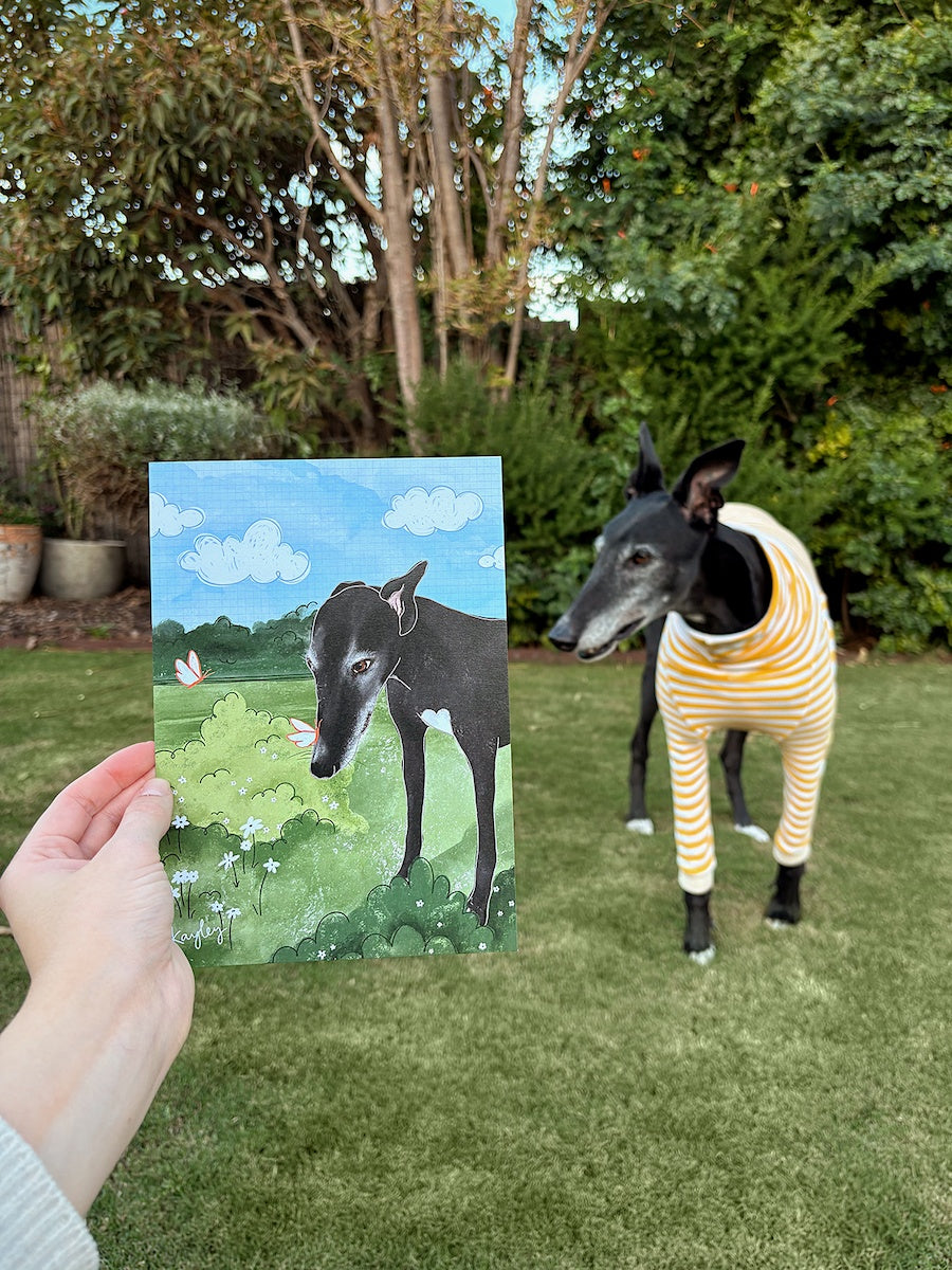 Chilli, a black greyhound, stands behind a drawing of him in the garden