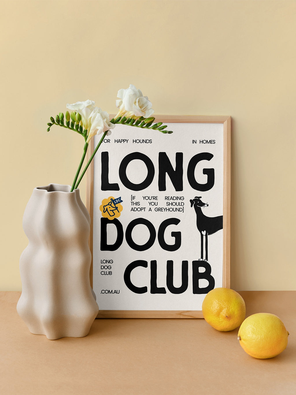 Framed 'Long Dog Club' poster on a table with a vase and lemons.