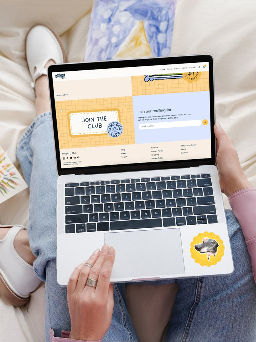 Person using a laptop with a website on the screen, sitting on a couch. A greyhound sticker is on the side.