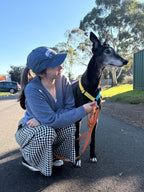 A woman crouches outdoors next to Chilli, a black greyhound. Kayley is wearing a blue cap, a blue cardigan, and black-and-white gingham pants.