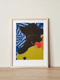 A framed screenprint of a black dog resting on a cuddlepillar toy. The portrait is of Elliott Fox's greyhound, Adam Barker. 
