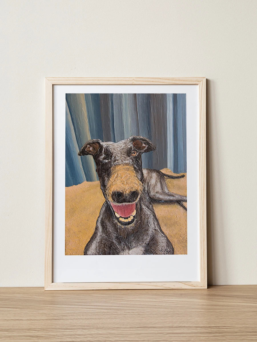 A framed pet portrait print of Gracie the Greyhound painted by Elliott Fox. Gracie has one eye and is smiling at the camera with dirt on her nose.