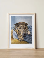 A framed pet portrait print of Hannah the greyhound resting on blankets, displayed in a wooden frame. Painted by Elliott Fox.