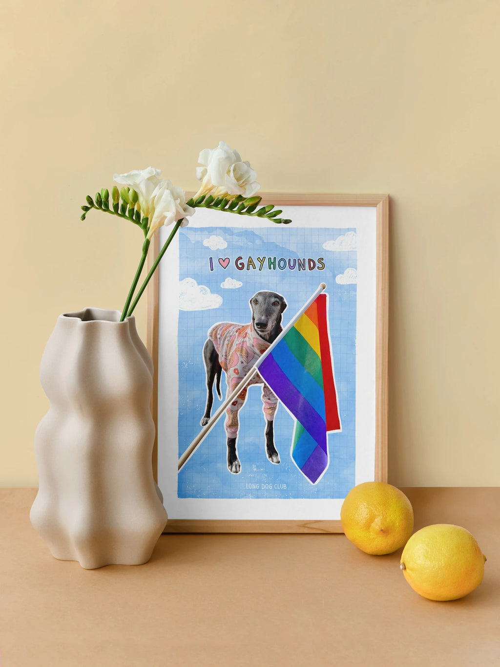 Framed artwork of a dog with a rainbow flag and 'I ❤️ Gayhounds' text, next to a vase with flowers and lemons on a beige surface.
