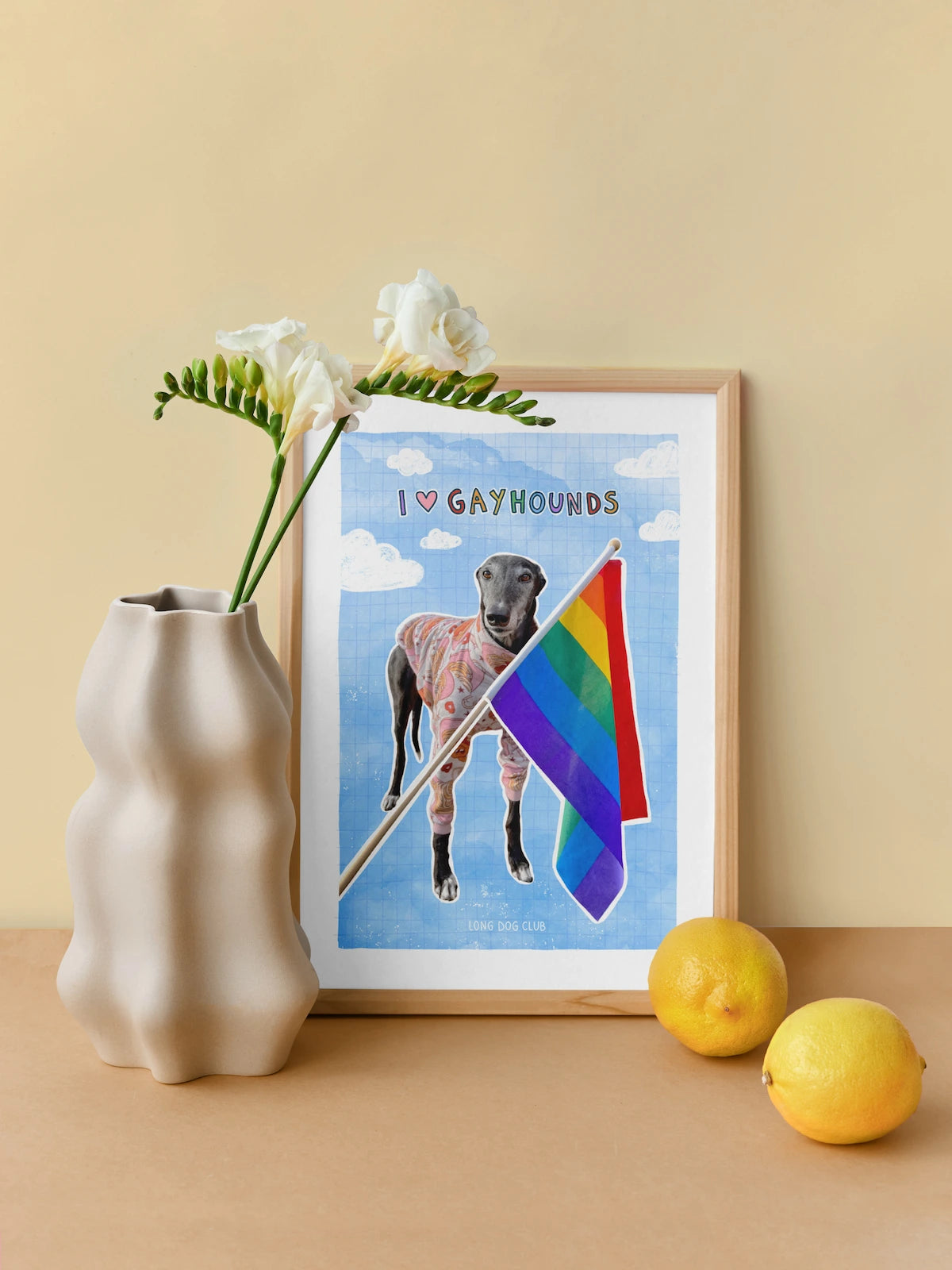 Framed artwork of a dog with a rainbow flag and 'I ❤️ Gayhounds' text, next to a vase with flowers and lemons on a beige surface.