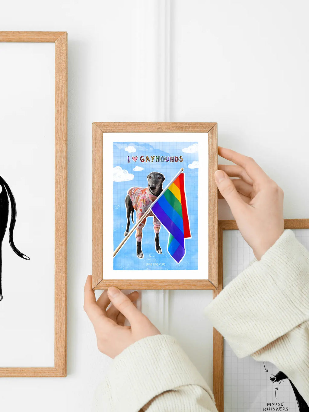 Person holding a framed print of a greyhound dog with a rainbow flag against a white wall.