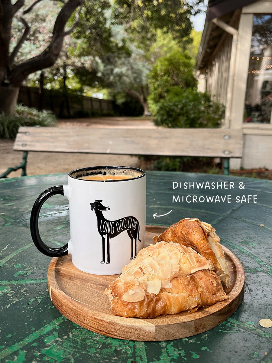 Ceramic greyhound mug sits at a cafe table outside next to a croissant. Text reads "dishwasher & microwave safe”