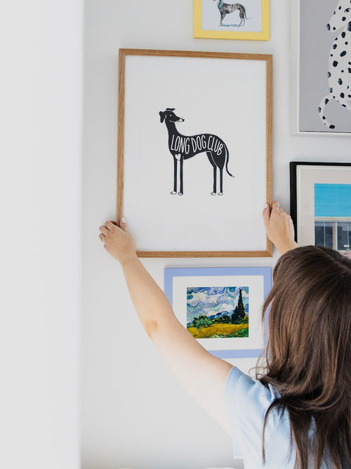 Woman is hanging a greyhound illustration print on a gallery wall of dog art