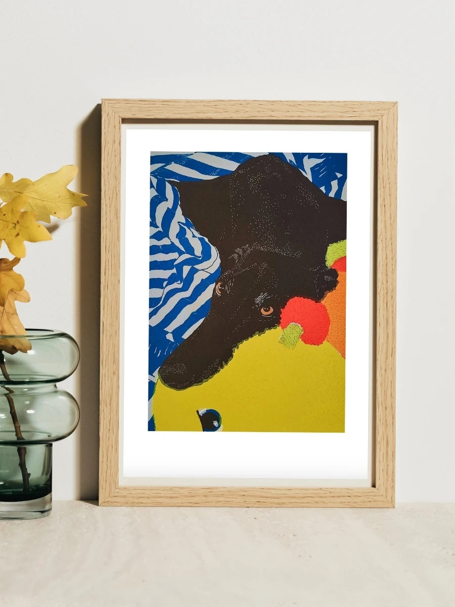 A framed screenprint of a black greyhound called Adam Barker cuddling a soft toy. Painted by Elliott Fox with the art print sold at Long Dog Club to raise money for greyhound welfare.