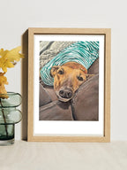 A framed pet portrait print by Elliott Fox leaning against a light wall on a tabletop, next to a glass vase with yellow flowers. The artwork shows a fawn greyhound wearing a striped shirt looking at the camera.