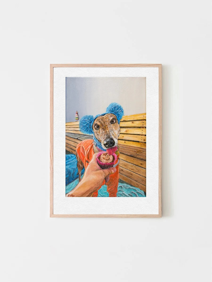 A framed art print by Elliott Fox featuring a bright portrait of a greyhound called Diva licking a cupcake. She wears a colourful outfit as looks very adorable.